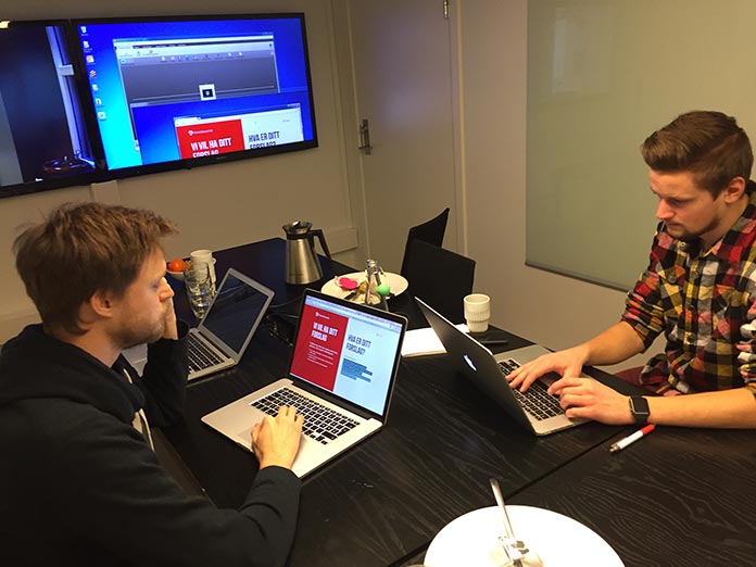 Two men sitting at a table and working on laptops, with a large screen in the background to display what they are collaborating on